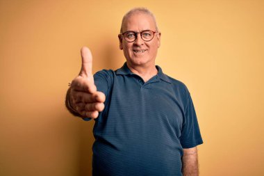 Orta yaşlı yakışıklı beyaz adam sarı arka planda günlük polo ve gözlük takıyor gülümseyerek dostça selamlaşıp selamlaşıyorlar. Başarılı bir iş..