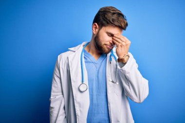 Sakallı, mavi gözlü, beyaz önlüklü, steteskop takan, burnu ve gözleri ovuşturan yorgun, yorgun ve başı ağrıyan genç sarışın doktor. Stres ve hüsran kavramı.