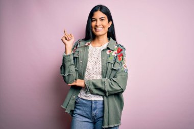 Pembe arka planda havalı bir ceket giyen, yüzünde kocaman bir gülümseme olan, elleri ve parmaklarıyla kameraya bakan genç ve güzel İspanyol bir moda kadını..