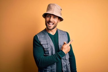 Tatilde sakallı yakışıklı bir turist sarı arka planda kaşif şapkası takıyor. Gülümsüyor ve gülümsüyor. Yüzünde mutlu ve doğal bir ifade var.