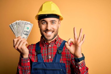 Güvenlik kaskı takan genç müteahhit sarı arkaplan üzerinde dolar tutuyor. Tamam işareti yapıyor, mükemmel bir sembol.