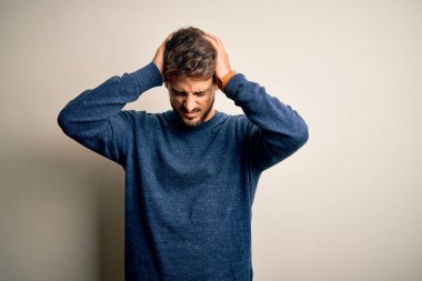 Sakallı, sıradan bir kazak giyen, beyaz arka planda dikilen, baş ağrısı çeken, çaresiz ve stresli genç bir adam. Çünkü acı ve migren. Ellerini başının üstüne koy..