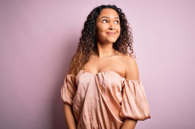 Kıvırcık saçlı, rahat tişörtlü pembe arka planda durup gülümseyen ve gözlerini kaçıran genç bir kadın..
