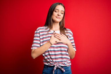 Genç, güzel, esmer, sıradan çizgili tişört giyen, kırmızı arka planda duran, elleri göğsünde, gözleri kapalı ve yüzünde minnet dolu bir jest yapan bir kadın. Sağlık kavramı.