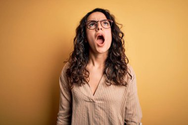 Kıvırcık saçlı, çizgili tişörtlü, sarı arka planda gözlüklü kızgın, sinirli ve öfkeli bir kadın. Öfkeyle bağırıyor. Öfke ve agresif kavram.
