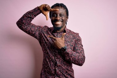 Genç, yakışıklı, Afro-Amerikalı bir adam pembe arka planda dikilmiş gülümsüyor ve elleri ve parmakları mutlu yüzlü bir çerçeve yapıyor. Yaratıcılık ve fotoğrafçılık kavramı.