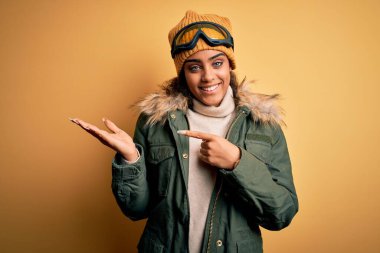 Genç Afrikalı Afro-Amerikan kayakçı kız kar sporu giysisi ve kayak gözlüğü takıyor kameraya hayret ediyor ve gülümsüyor..