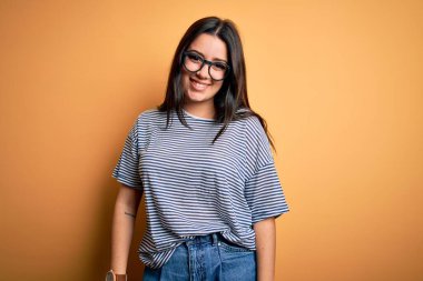 Genç esmer kadın gözlüklü ve donanma tişörtlü sarı izole arka planda mutlu ve havalı bir gülümsemesi var. Şanslı kişi..