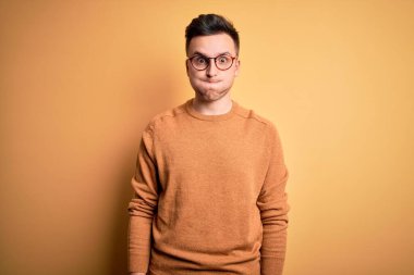 Genç, beyaz, gözlük takan yakışıklı bir adam ve sarı arka planda komik yüzlü, pofuduk yanaklı, sıradan bir kazak giyiyor. Havayla şişmiş ağız, çılgın bir ifade..