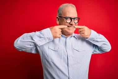 Orta yaşlı yakışıklı beyaz adam kırmızı arka planda günlük çizgili gömlek ve gözlük takıyor. Gülümsüyor, parmaklarını ve ağzını göstererek gülümsüyor. Diş sağlığı konsepti.