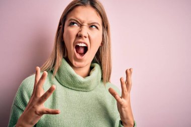 Pembe izole edilmiş arka plan üzerine kışlık yün kazak giyen genç, güzel sarışın kadın sinirli bir şekilde bağırıyor ve agresif bir ifade ve kolla bağırıyor. Hayal kırıklığı konsepti.