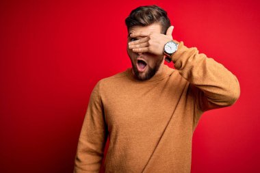 Sakallı ve mavi gözlü genç bir adam kırmızı arka planda sıradan bir süveter giyiyor. Şok olmuş bir şekilde yüzünü ve gözlerini kapıyor. Utanmış bir ifadeyle parmaklarının arasından bakıyor..