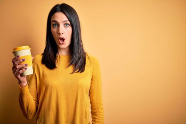 Mavi gözlü, elinde kahve fincanı olan genç bir kadın sarı arka planda duruyor. Şaşırmış bir yüzle korkmuş ve korku ifadesiyle heyecanlanmış.
