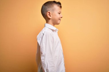 Mavi gözlü, sarı arka planda zarif beyaz gömlek giyen, rahat bir görünüm takınan, kendine güvenen gülümseyen küçük beyaz çocuk..