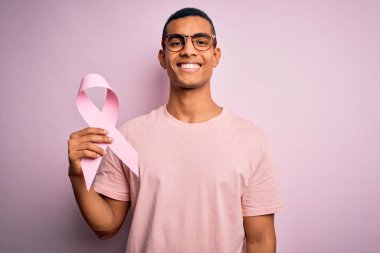 Genç, yakışıklı, Afro-Amerikan bir adam pembe kanser kurdelesini izole edilmiş arka planda saklıyormuş. Mutlu bir yüzü varmış ve gülümsüyormuş.
