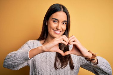 Sarı arka planda beyaz süveter giyen genç, güzel esmer kadın. Aşkla gülümsüyor. Elleriyle kalp sembolü yapıyor. Romantik konsept.