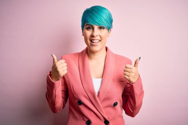 Pembe arka plan başarı işareti üzerine ceket giyen, mavi saçlı genç ve güzel bir iş kadını. Elleriyle olumlu jest yapıyor. Gülümsüyor ve mutlu. Neşeli ifade ve kazanan jest.