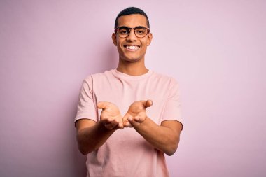 Yakışıklı Afro-Amerikalı adam pembe arka planda günlük tişört ve gözlük takıyor avuçlarını birbirine değdirip gülümsüyor ya da el hareketi yapıyor. Tutun ve koruyun