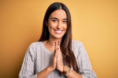 Sarı arka plan üzerine beyaz süveter giyen genç, güzel esmer kadın ellerinin birarada dua ederek af diliyor kendinden emin bir şekilde gülümsüyor..