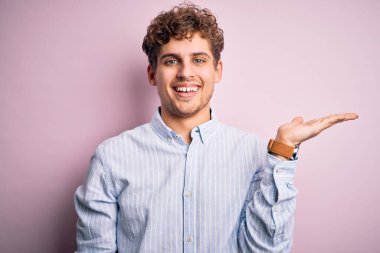 Sarışın, kıvırcık saçlı, beyaz arka planda çizgili gömlek giyen neşeli bir şekilde gülümseyen ve avuç içi kameraya bakan genç bir adam..