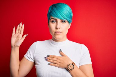 Mavi saçlı genç bir kadın kırmızı arka planda rahat bir tişört giyiyor göğsü ve avucu açık yemin ediyor sadakat yemini ediyor.