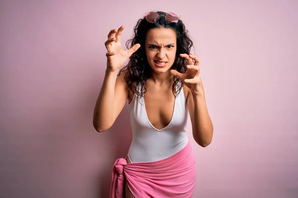 Beautiful woman with curly hair on vacation wearing white swimsuit over pink background Shouting frustrated with rage, hands trying to strangle, yelling mad