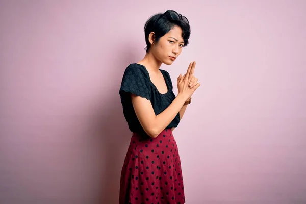 Young beautiful asian girl wearing casual dress standing over isolated pink background Holding symbolic gun with hand gesture, playing killing shooting weapons, angry face