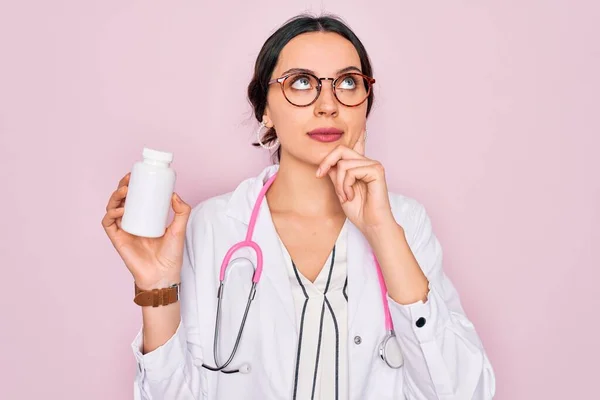 Young beautiful doctor woman with blue eyes wearing stethoscope holding medicine pills serious face thinking about question, very confused idea