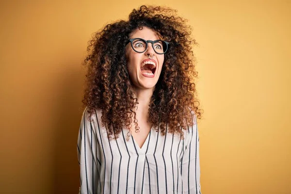 Young beautiful woman with curly hair and piercing wearing striped shirt and glasses angry and mad screaming frustrated and furious, shouting with anger. Rage and aggressive concept.