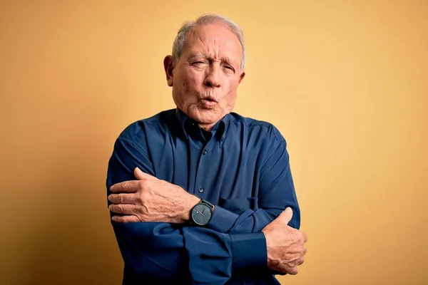 Grey haired senior man wearing casual blue shirt standing over yellow background shaking and freezing for winter cold with sad and shock expression on face