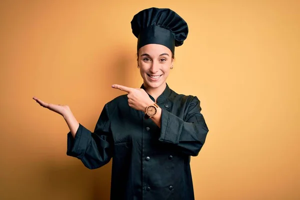 年轻美丽的厨师女士身穿炊事服 头戴一顶帽子 站在黄色的背景上 满脸诧异地看着摄像机 手牵着手 指指点点着镜头 — 图库照片