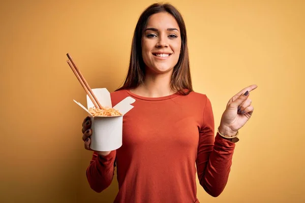 年轻美丽的黑发女人用筷子吃着外卖的面条 她很高兴地将手和手指指向旁边 — 图库照片