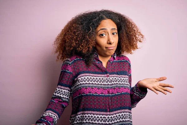 Young african american woman with afro hair wearing colorful shirt over pink background clueless and confused expression with arms and hands raised. Doubt concept.