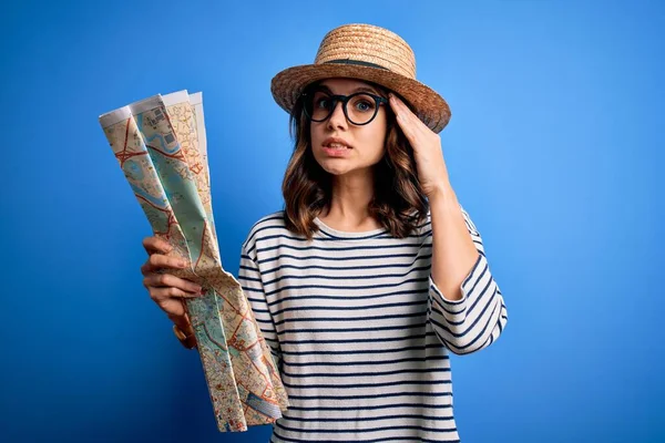 Young blonde tourist girl wearing a hat looking at trip destination city map over blue background stressed with hand on head, shocked with shame and surprise face, angry and frustrated. Fear and upset for mistake.