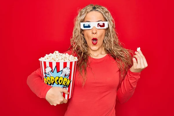 Young beautiful blonde woman watching movie using 3d glasses eating popcorn scared and amazed with open mouth for surprise, disbelief face