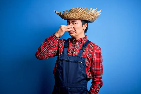 Young handsome chinese farmer man wearing apron and straw hat over blue background smelling something stinky and disgusting, intolerable smell, holding breath with fingers on nose. Bad smell