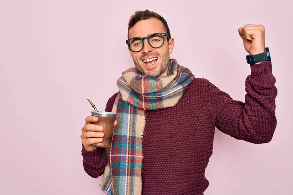 Young handsome man with blue eyes drinking cup of hot mate tea over pink background screaming proud and celebrating victory and success very excited, cheering emotion