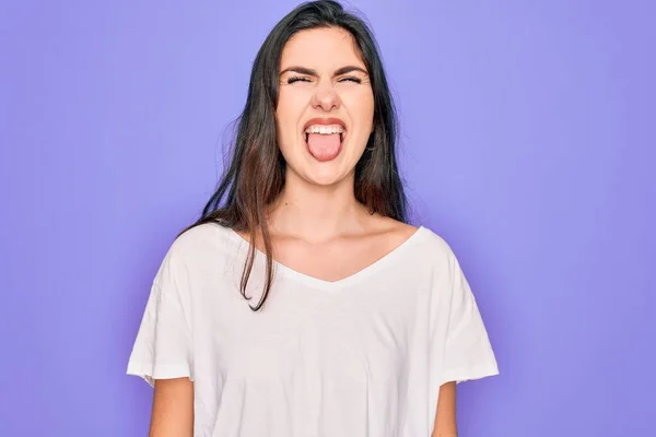 紫の背景にカジュアルな白いTシャツを着た若い美しいブルネットの女性が面白い表情で幸せ舌を突き出している 感情概念 — ストック写真