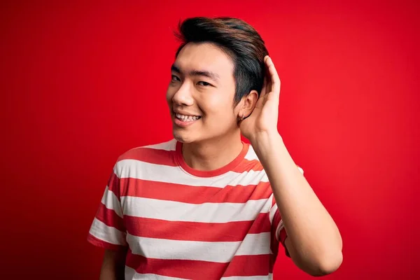 Young handsome chinese man wearing casual striped t-shirt standing over red background smiling with hand over ear listening an hearing to rumor or gossip. Deafness concept.