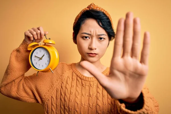 年轻美丽的中国女人 在孤立的黄色背景上握着老式闹钟 张开手 做了一个严肃而自信的手势 做了一个防御手势 — 图库照片