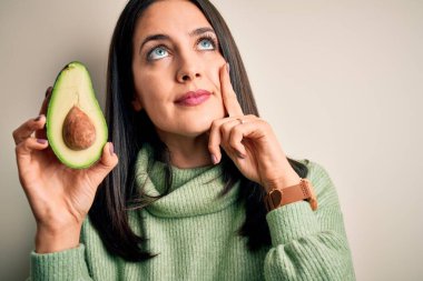 Mavi gözlü, orta sağlıklı avokadoyu soyutlanmış beyaz arka planda tutan genç bir kadın. Ciddi bir yüz. Soru sormayı düşünüyor.