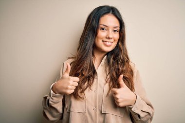 Beyaz arka plan başarısı tabelasının üzerinde duran rahat gömlekli genç, güzel esmer kadın. Elleriyle olumlu jest yapıyor. Gülümsüyor ve mutlu. Neşeli ifade ve kazanan jest.