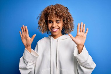 Güzel Afro-Amerikan sportif kadın mavi arka plan üzerine günlük kazak giyiyor ve parmaklarını on numarayı gösterirken kendinden emin ve mutlu bir şekilde gülümsüyor..