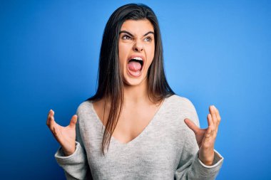 Genç, güzel, esmer bir kadın mavi arka planda dikilmiş deli deli deli bağırıyor ve agresif bir ifadeyle ve kollarıyla bağırıyor. Hayal kırıklığı konsepti.