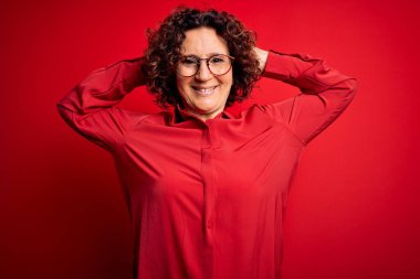 Orta yaşlı güzel kıvırcık saçlı kadın kırmızı arka planda günlük gömlek ve gözlük takıyor rahatlıyor ve esniyor, kolları ve elleri başının arkasında ve boynunun arkasında gülümsüyor.