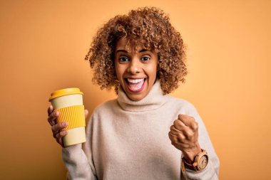 Genç Afro-Amerikan kıvırcık kadın sarı arka planda bir fincan kahve içerken gururla bağırıyor ve zaferi ve başarıyı kutluyor.