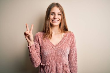 Genç, güzel, kızıl saçlı, pembe bir süveter giyen, izole edilmiş beyaz arka planda iki numarayı işaret eden, kendinden emin ve mutlu bir şekilde gülümseyen bir kadın..