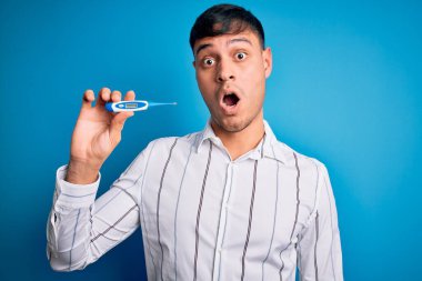 Tıbbi termometreyi mavi arkaplanda tutan genç İspanyol adam şaşırmış bir yüzle korkmuş ve korku ifadesinden heyecanlanmış.
