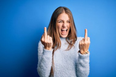 Young beautiful redhead woman wearing casual sweater over isolated blue background Showing middle finger doing fuck you bad expression, provocation and rude attitude. Screaming excited