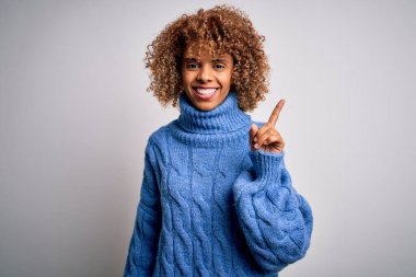 Beyaz arka plan üzerine balıkçı yaka kazak giyen genç ve güzel bir Afro-Amerikalı kadın. Yüzünde kocaman bir gülümseme, kameraya el parmağı ile bakıyor..